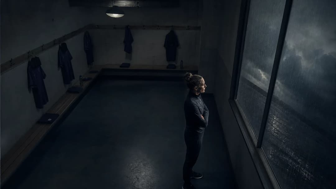 A female football manager in an empty locker room deep in thought, under dim lighting and heavy rain outside.