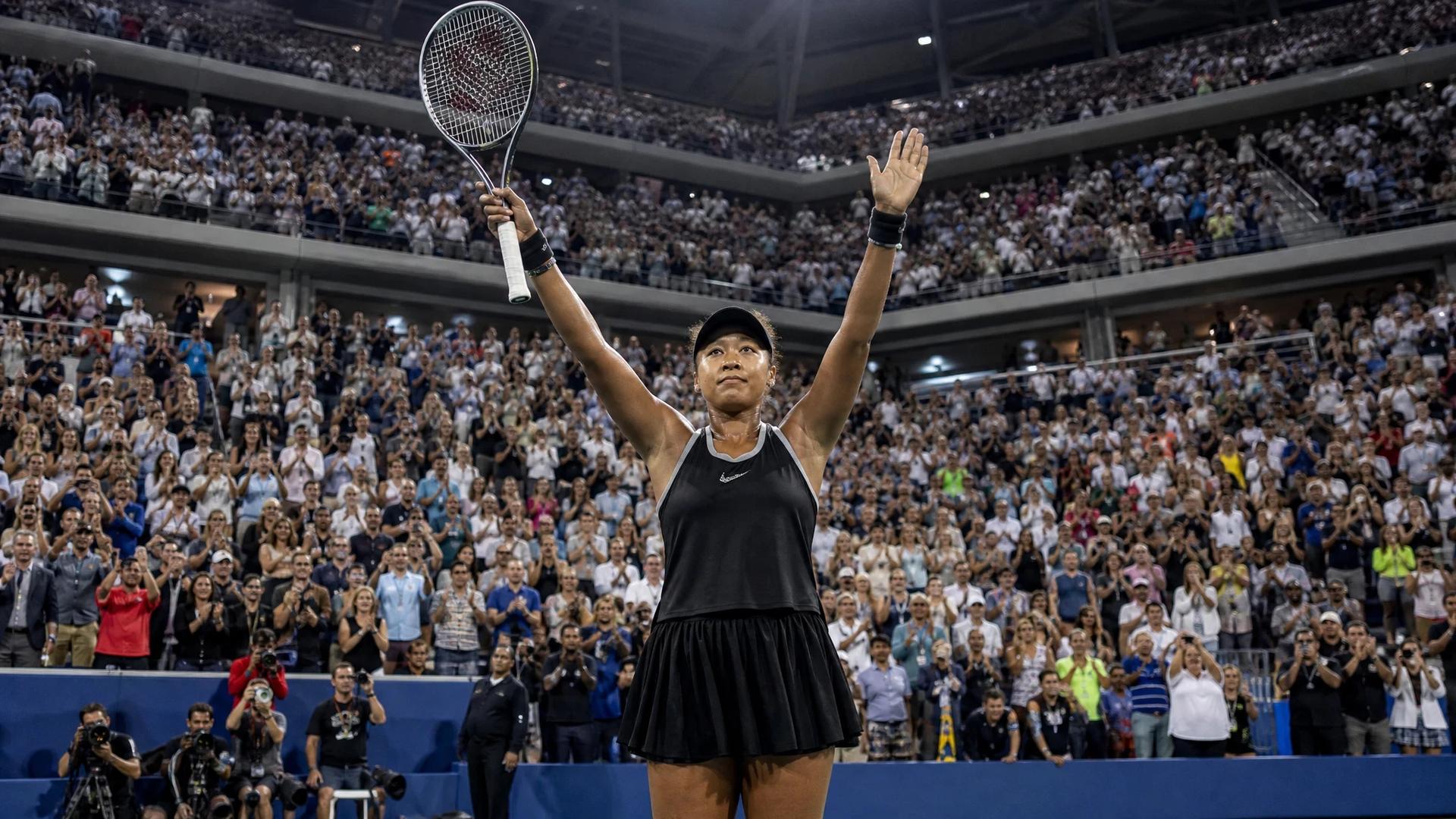 Naomi Osaka celebrating after a match, surrounded by a cheering crowd at the US Open.