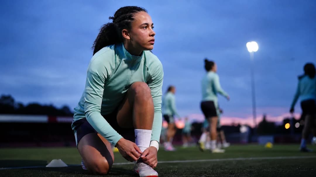 A close-up of Lisa Baum preparing for a football match at dusk with teammates blurred in the background.