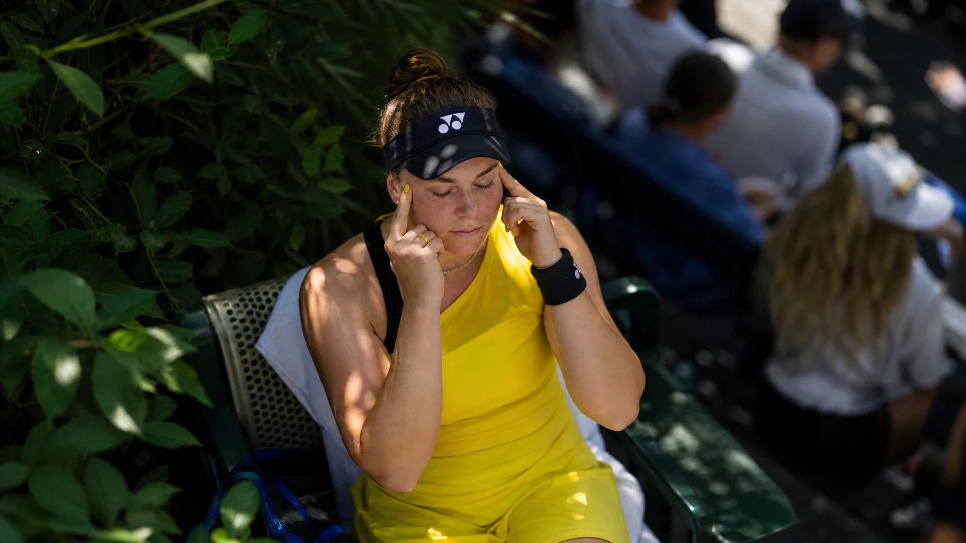 A bird's-eye view of Linda Nosková sitting on a bench with her fingers in her ears, surrounded by greenery.