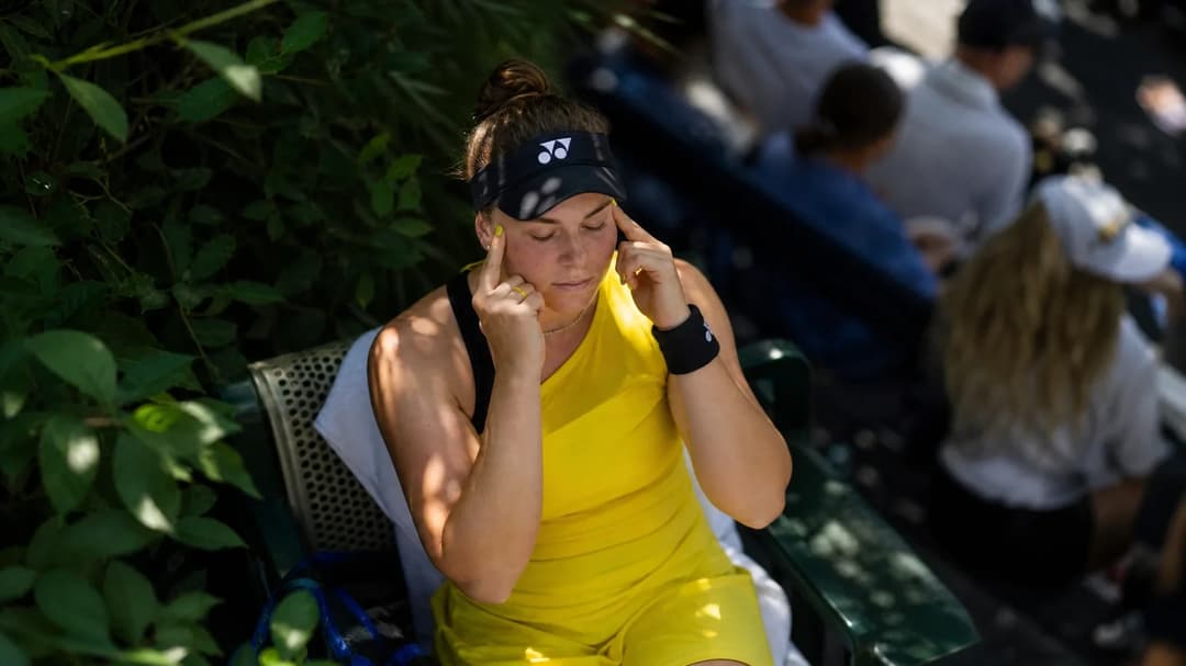 A bird's-eye view of Linda Nosková sitting on a bench with her fingers in her ears, surrounded by greenery.