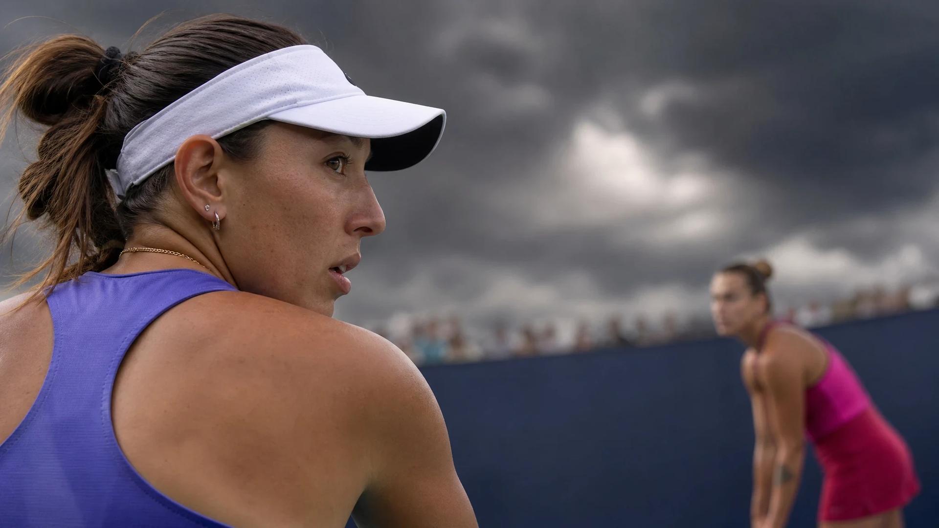 Jessica Pegula preparing to serve during her match at the US Open, with Aryna Sabalenka blurred in the background.