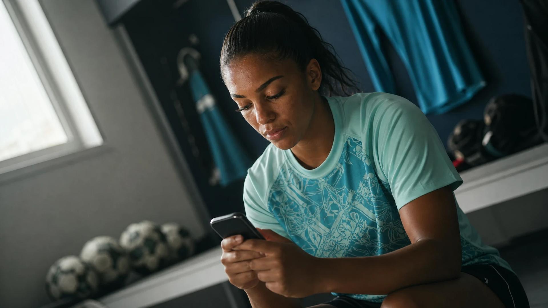 A female footballer sits on a bench in a training room, focused on her smartphone.