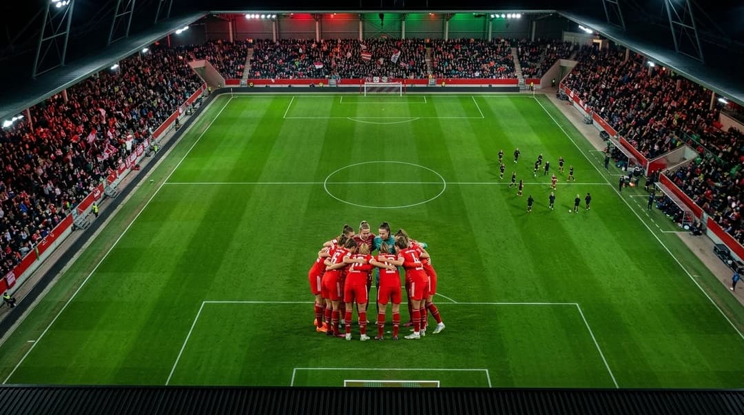 Aerial view of Bayern Munich women's team in a strategic huddle on a football pitch, surrounded by a cheering crowd.