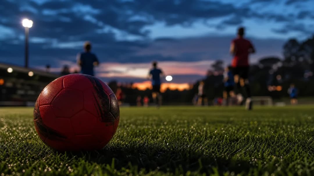 A close-up of a football on grass with blurred players in the background during dusk.